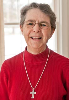 Elderly woman with short gray hair and glasses, wearing a red sweater and a silver cross necklace, smiling in front of a window decorated for Spring Break. Saint Joseph's College of Maine