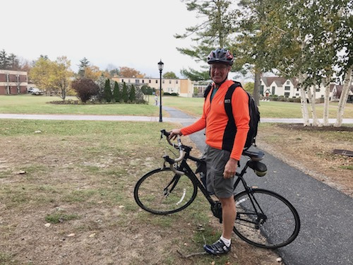 A person wearing an orange jacket and helmet stands next to a black bicycle on a path in a residential area, proudly displaying their Sustainable Commuting Award from Saint Joseph’s College as part of the Way 2 GO MAINE Challenge. Saint Joseph's College of Maine