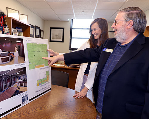 Two people standing by a table, pointing at architectural plans on a large board. They appear to be steering the discussion towards new design features shown on the display. Saint Joseph's College of Maine