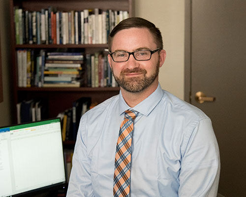 A man with short hair and glasses, dressed in a light blue shirt and orange plaid tie, sits in an office beside a computer and bookshelves, surrounded by legal texts that signify his commitment to justice. Saint Joseph's College of Maine