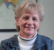 A woman with short-blonde hair smiles, wearing a grey blazer over a white turtleneck. Behind her, a world map hints at the global reach of the online radiology program she's discussing. Saint Joseph's College of Maine