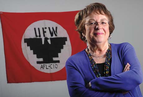 A woman wearing a blue top stands with her arms crossed in front of a red flag displaying the UFW and AFL-CIO logos, embodying the spirit of "Sí Se Puede. Saint Joseph's College of Maine