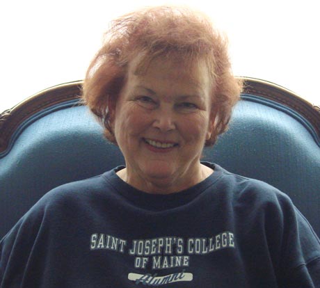 Cornelia Baker, with her short, curly red hair, sits in a blue upholstered chair, smiling. She is wearing a navy sweatshirt that proudly reads "Saint Joseph's College of Maine Alumni. Saint Joseph's College of Maine