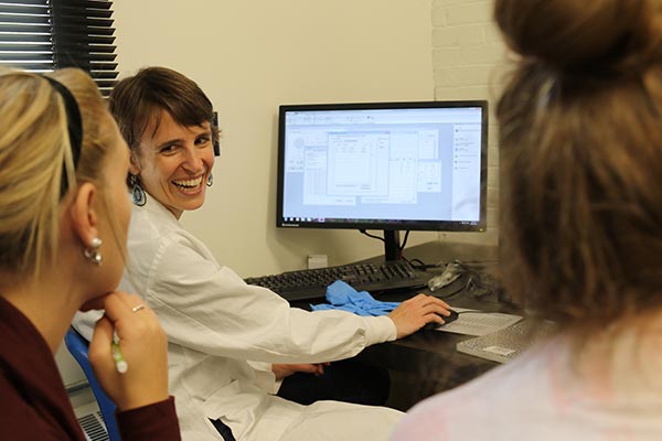 A woman in a lab coat smiles while sitting at a computer, demonstrating something on the screen to two others, embodying the spirit of science and serving through knowledge sharing. Saint Joseph's College of Maine