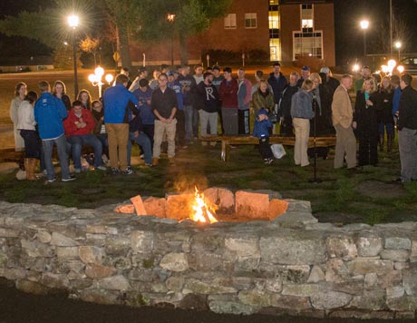 A group of people gather around an outdoor fire pit at night, with some seated on benches and others standing. The warm glow acts as a welcomed light, while nearby buildings and streetlights provide additional lighting, creating a cozy ambiance that feels like an extension of thoughtful home decor. Saint Joseph's College of Maine