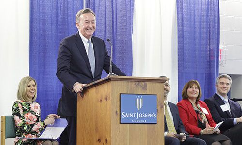 A man speaks passionately at a podium labeled "Saint Joseph's College," while five seated individuals listen attentively against a backdrop of blue curtains, highlighting the significance of the Harold Alfond Foundation Challenge Grant to the institution. Saint Joseph's College of Maine