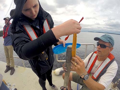 Two individuals on a boat, each wearing life vests, are examining and handling scientific equipment while a third person stands nearby. They appear to be involved in field research on water, supported by the National Science Foundation Funding as part of the SJC Science Scholars Program. Saint Joseph's College of Maine