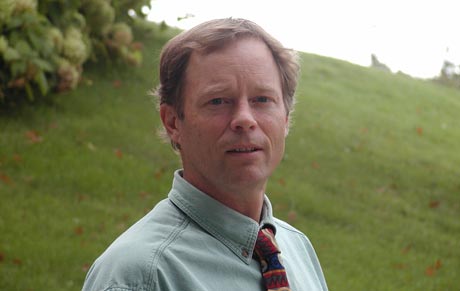 A man with short hair, wearing a green collared shirt and colorful tie, stands outdoors with greenery in the background, looking like a distinguished professor ready to discuss his latest geology research grant. Saint Joseph's College of Maine