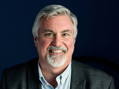 Mark Swann, a respected social service leader with gray hair and a beard, wearing a suit jacket and striped shirt, smiles against a dark blue background. Saint Joseph's College of Maine