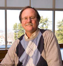 A man with glasses and a mustache is wearing a sweater with a diamond pattern, sitting indoors in front of a window with trees outside, reading new books. Saint Joseph's College of Maine