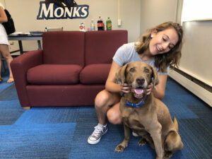 A woman kneels on the floor next to a brown dog, smiling and holding its head. They are in a room with a red couch and a table with drinks; "Monks" is written on the wall in the background. It's as if they are celebrating Yappy Hour, enjoying each other's company. Saint Joseph's College of Maine