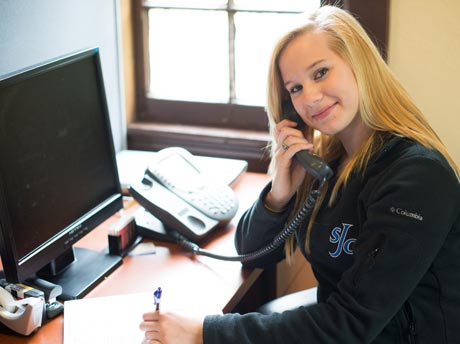 A woman with long blonde hair is sitting at her desk, talking on the phone, and writing on paper during the Fall Phonathon. A computer monitor and telephone are also on the desk. She is smiling brightly and looking at the camera, ready to connect with thousands of potential donors. Saint Joseph's College of Maine