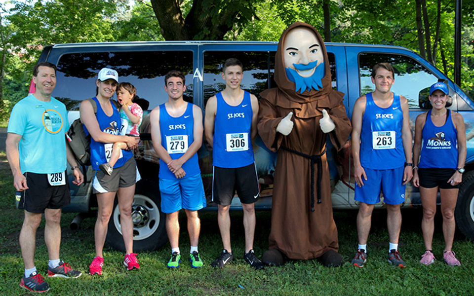 A group of people in athletic wear stand outdoors next to a large mascot in a monk costume. They are in front of a van, participating in the Westbrook Strong 5K event, likely part of raising awareness for the Rairdon Nursing Scholarship. Saint Joseph's College of Maine