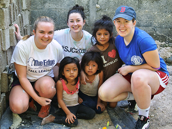 Three women and three children, embodying the spirit of serving the Common Good, pose for a photo in front of a partially constructed or damaged building. The women are squatting, and the children are sitting on the ground. Saint Joseph's College of Maine