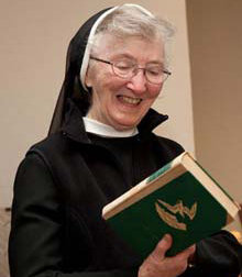 Sister Mary, an elderly woman in a nun's habit, smiles while holding and looking at a book with a green cover, enjoying her quiet moment of reflection in retirement. Saint Joseph's College of Maine