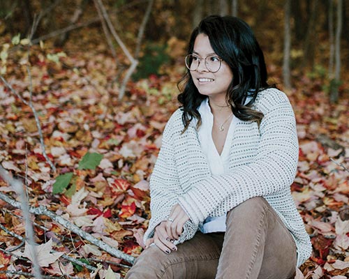 A woman with glasses and a white sweater sits on autumn leaves in a forest, looking off to the side as if finding inspiration for her book, "Breaking Through the Silence. Saint Joseph's College of Maine