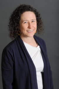 Counseling Center 1 A woman with curly brown hair, wearing a navy blue cardigan over a white top and large brown earrings, smiles while standing against a plain gray background. Saint Joseph's College of Maine