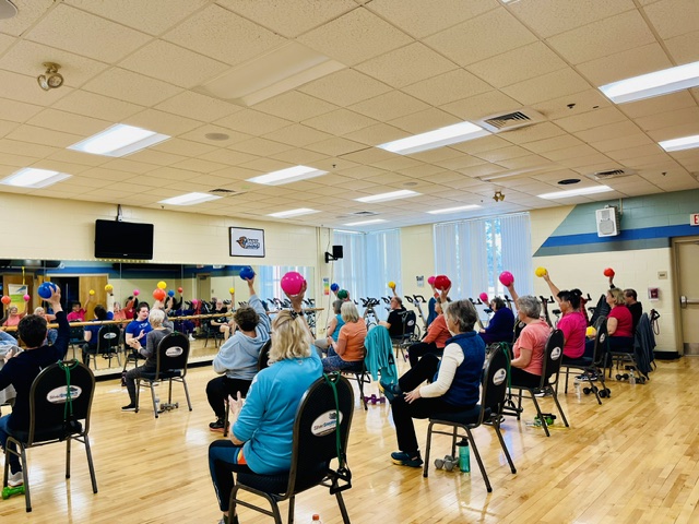 A group of adults participate in a SilverSneakers fitness class, sitting on chairs with colorful balloons attached. The gym features wooden floors, mirrors, and large windows that fill the space with natural light. Saint Joseph's College of Maine