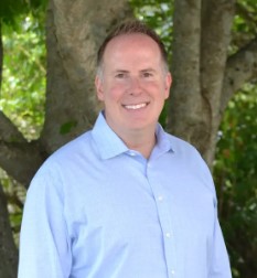 A man in a light blue button-up shirt stands smiling in front of a tree with green leaves in the background. Saint Joseph's College of Maine