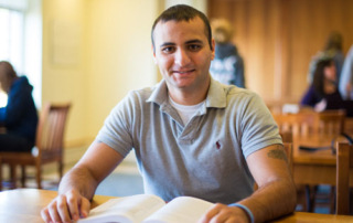 Boots &Amp; Books 1 A man sits at a library table with an open book in front of him, smiling at the camera. People in the background, immersed in their own books, are also engaged in studying. His worn boots peek out from under the table, suggesting he’s been here for a while. Saint Joseph's College of Maine