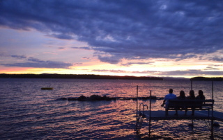 College Welcomes Home Hundreds Of Alumni For Reunion Weekend 5 Four alumni sit on a bench at the end of a dock, overlooking a lake during a colorful sunset with purple and orange hues in the sky, reminiscing about their college reunion. Saint Joseph's College of Maine