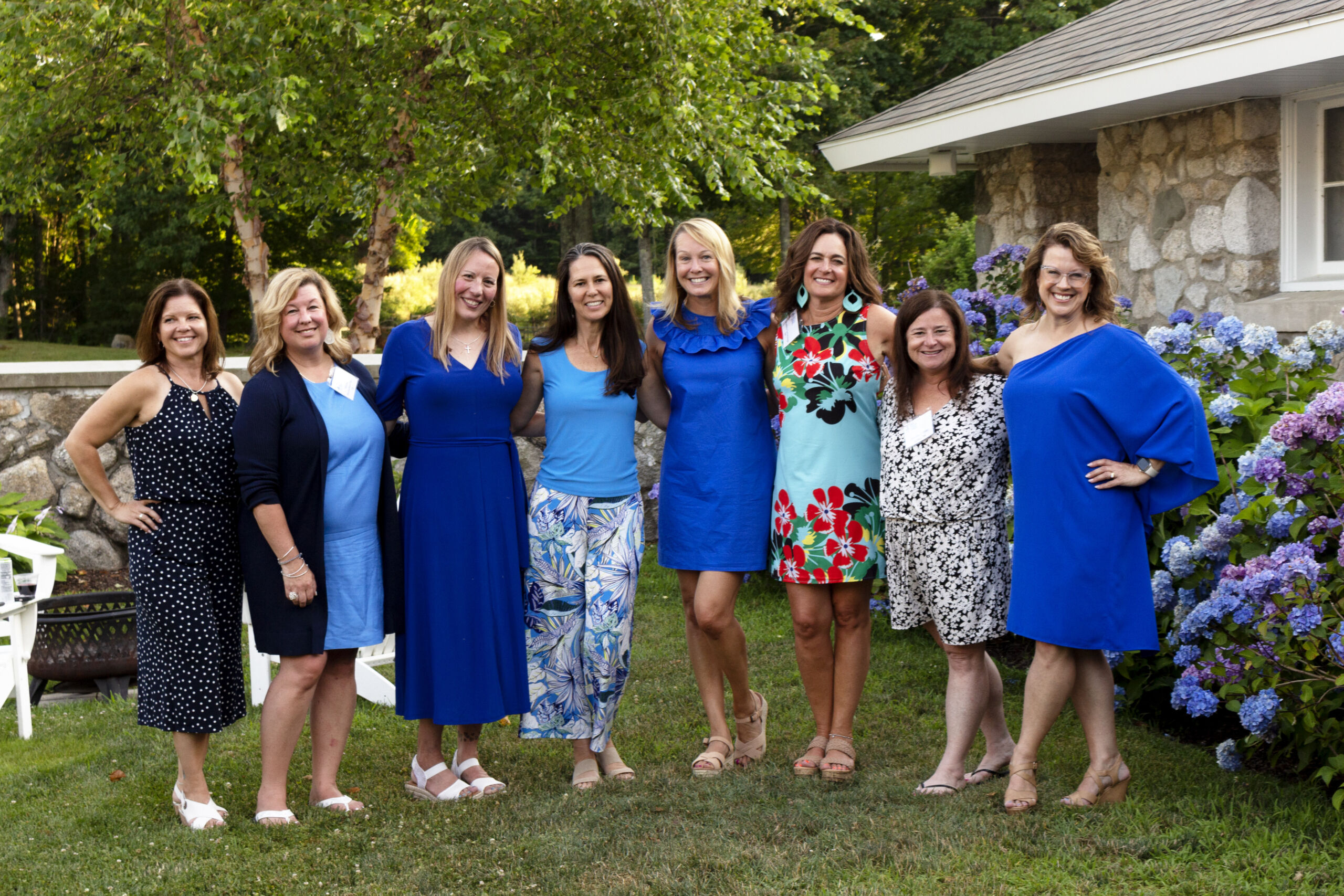 Alumni 1 A group of eight women standing on grass in a garden, smiling, with some wearing blue outfits and others in floral patterns. A stone building and hydrangea bushes are in the background. Saint Joseph's College of Maine