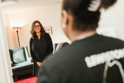 Counseling Center 1 A woman in glasses stands and smiles in a living room, facing another person whose back is to the camera, suggesting a warm and welcoming atmosphere like that found at a counseling center. Saint Joseph's College of Maine