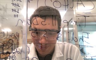 Looking For A Cure 5 A person wearing glasses and a lab coat is looking intently through a glass board covered with various handwritten notes and equations. Saint Joseph's College of Maine