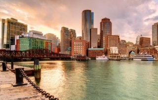 The Case For The Regional Alumni Chapter 9 Waterfront cityscape featuring high-rise buildings and boats docked at the harbor. A chain barrier lines the stone pathway along the water, where a regional alumni chapter gathers for their annual reunion. The sky shows hints of sunset or sunrise colors, adding to the picturesque scene. Saint Joseph's College of Maine