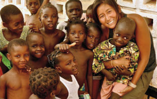 Living The Mission By Serving Others 1 A smiling woman, living the mission, is surrounded by a group of children in an outdoor setting. She is holding one of the children, and all are looking at the camera, serving others with warmth and joy. Saint Joseph's College of Maine