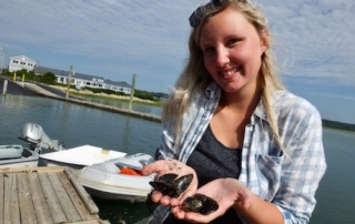 Brooke Murch ’18 Conducts Scientific Sensor Research At Wells National Estuarine Research Reserve 9 A smiling Brooke Murch, holding two mussels, stands on a dock with boats and buildings in the background, likely near the Wells National Estuarine Research Reserve. Saint Joseph's College of Maine