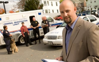 Beyond The ‘Cop Shop’ 9 A man in a beige suit holds a clipboard outside, while several police officers and a police dog stand near a "Criminal Investigation Unit" vehicle and police car beyond the cop shop in the background. Saint Joseph's College of Maine