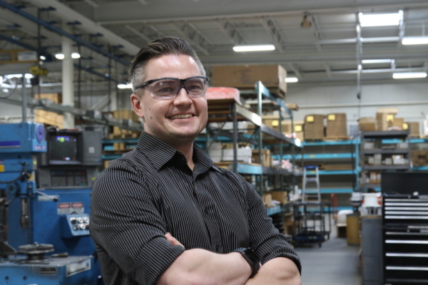 Leading In A World Of Tension 1 Bryon Williams poses, wearing safety glasses, on the work floor in front of shelves of tools