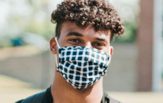 A person with curly hair wearing a checkered face mask and a black shirt is standing outdoors with a backpack. Saint Joseph's College of Maine