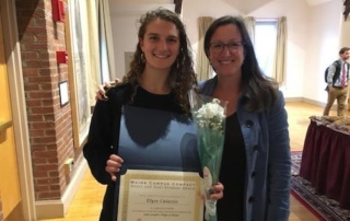 Maine Campus Compact Honors Elyse Caiazzo ’18 With The Heart &Amp; Soul Student Award And The Sjc Ecoreps With The President’s Campus Leadership Award 2 Two women indoors, smiling. The woman on the left is holding a certificate and a flower bouquet, having just received the Heart & Soul Student Award. The woman on the right stands beside her with her arm around her shoulder. Saint Joseph's College of Maine