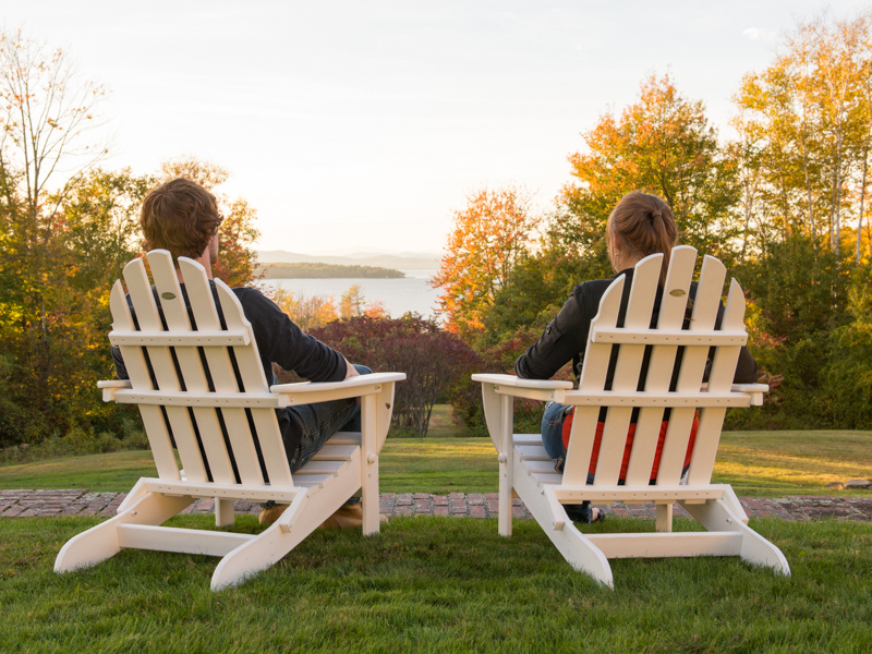 Two people sit in white Adirondack chairs on a grassy lawn, facing a lake surrounded by trees with autumn foliage at Saint Joseph's College of Maine. Saint Joseph's College of Maine