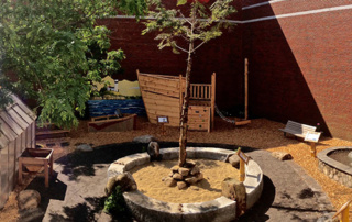 Unbound 5 An unbounded outdoor play area features a central tree surrounded by a circular sandpit, wooden structures resembling a boat, and a bench against a brick wall. Saint Joseph's College of Maine