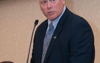 A man wearing a suit and tie speaks into a microphone at a podium, looking forward with determination as he begins Part 7 of his presentation. Saint Joseph's College of Maine