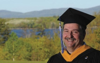 David Stamm ’11 In His Own Words 2 David Stamm, in his own words, stands proudly outdoors in graduation attire. He wears a black cap and gown with a gold-colored sash, against a scenic backdrop of trees and a shimmering lake. Saint Joseph's College of Maine