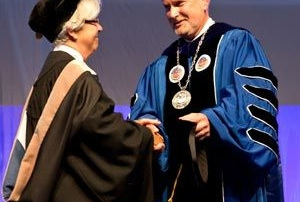 Saint Joseph’s Inaugurates President James Dlugos 2 At Saint Joseph's Inauguration, two individuals in academic regalia shake hands—one in a black cap and gown, the other, President James Dlugos, in a blue gown adorned with medals. Saint Joseph's College of Maine