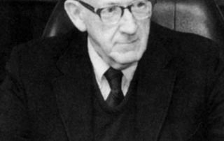 The Hart Of Campus 4 An older gentleman in a suit and tie sits at a desk with a pen in hand, looking thoughtfully to the side, embodying the wisdom that forms the Hart of Campus. Saint Joseph's College of Maine