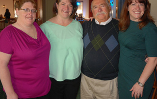 A Decade Of Building Florence Dussault’s Legacy 8 Four people stand close together, smiling at the camera. The group consists of three women and one man wearing a sweater with an argyle pattern. They appear to be in a warmly lit indoor setting, perhaps celebrating Florence Dussault’s Legacy. Saint Joseph's College of Maine