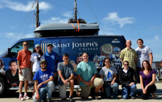 Students, Faculty Take Science On The Road With Ess 4 A group of eleven students and faculty members stands in front of a blue Saint Joseph’s College van with a boat on the roof, smiling at the camera. The van features the college's logo and various sports images. Saint Joseph's College of Maine
