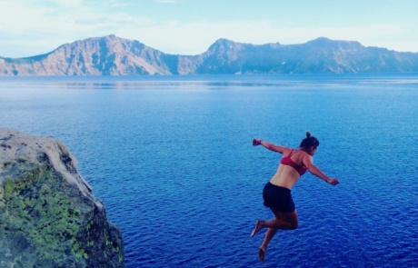 Crater Lake, Oregon A person in a red top and black shorts jumps off a cliff into a deep blue body of water with mountains in the background under a clear sky, experiencing pure trail magic. Saint Joseph's College of Maine