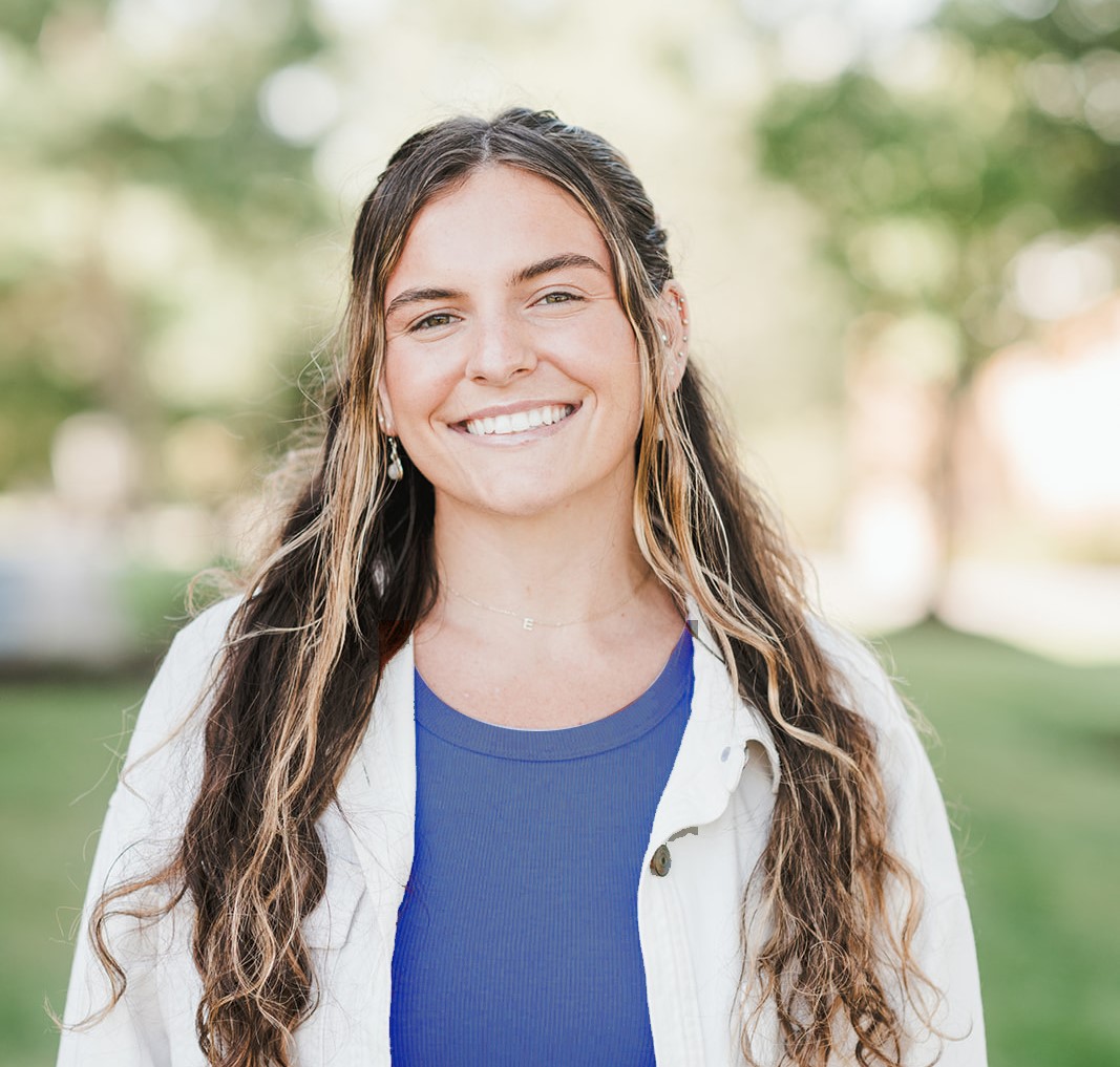 Emily Dion A young woman with long wavy hair, wearing a blue top and light jacket, smiles at the camera outdoors with greenery in the background. Saint Joseph's College of Maine