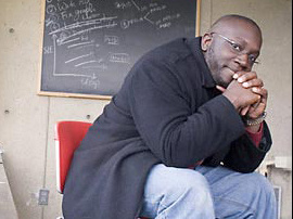 Erby Mitchell '98 Heads Admissions At Elite School And Admits President Obama’s Daughters 1 A person wearing glasses and a dark coat sits on a red chair in front of a chalkboard filled with notes and diagrams, resting their hands on their chin and looking directly at the camera, as if contemplating the secrets behind elite school admissions. Saint Joseph's College of Maine