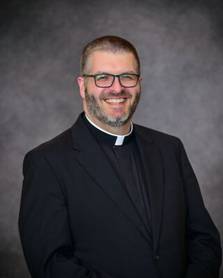 A man with short hair, a beard, and glasses, wearing a clerical collar and a black suit, smiles against a gray background. He serves as a chaplain at Saint Joseph's College in Maine. Saint Joseph's College of Maine