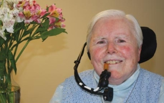 Sister Anne Fitzpatrick ’64, Rsm 1 Elderly Sister Anne Fitzpatrick with short white hair uses an assistive communication device. A vase of flowers is in the background. Saint Joseph's College of Maine