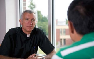 Talk - An Interview With Father Lou Phillips 10 Father Lou Phillips, dressed in a clerical shirt, listens attentively to another person in a green shirt during an interview inside a room with a large window. Saint Joseph's College of Maine