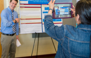 Spotlight On Research: Class Of 2015 4 A person points to a poster spotlighting the Class of 2015, while another individual photographs the scene using a tablet. Saint Joseph's College of Maine